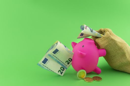 Reaping the rewards of financial success with a cute pink piggy bank, sack of money with a twenty Euro bill in the slot over a green background