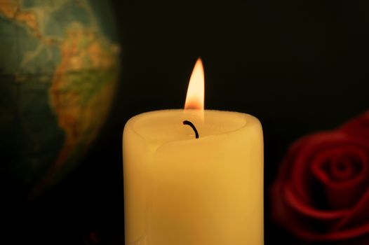 Single burning yellow candle in front of a globe next to red and yellow roses against dark background, concept of global disasters and deaths