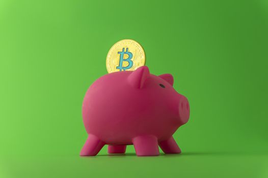Saving using Bitcoin concept with a virtual gold coin being inserted into a pink piggy bank over a green background