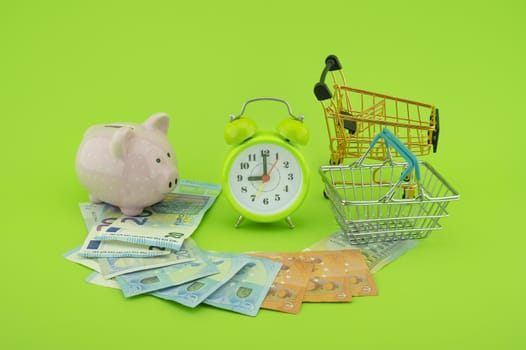 Cost of shopping or spending savings concept with banknotes scattered on a green background with a piggy bank, clock, small wire shopping basket and cart