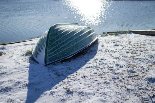 Small wooden dinghy or rowboat on a cold white frosty bank above a lake or river with sparkling sunshine reflecting on the water