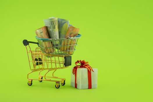 Miniature shopping cart filled with cash banknotes and a small gift box isolated on green background. Saving money for spending on gifts concept