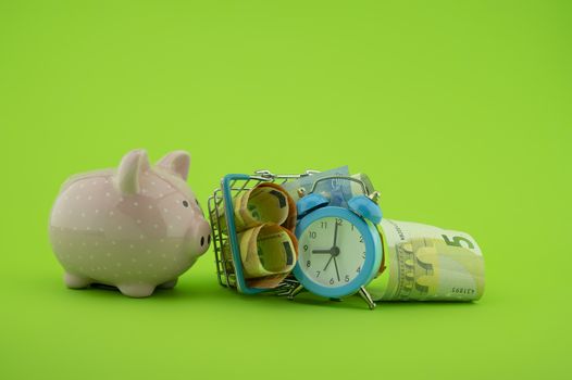 Shopping concept with ceramic piggy bank, basket filled with cash banknotes and alarm clock over a green background