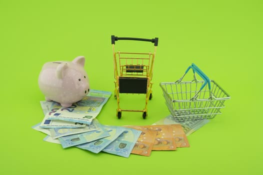 Cost of shopping or spending savings concept with banknotes scattered on a green background with a piggy bank, small wire shopping basket and cart