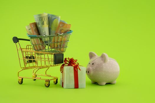 Miniature shopping cart filled with cash banknotes, next to a small gift box and ceramic piggy bank isolated on green background. Saving money for spending on gifts concept