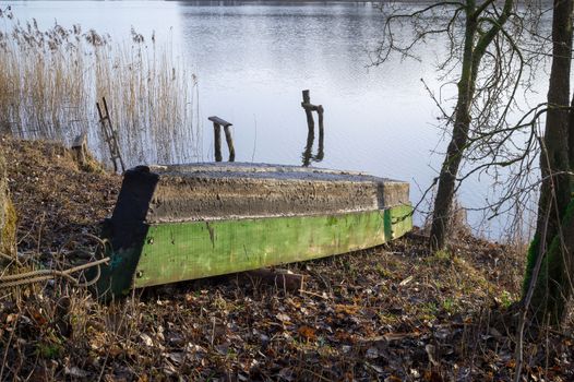 Green painted hull of an old wooden dinghy or rowboat pulled up on a lake shore standing up side down on the grass alongside calm water