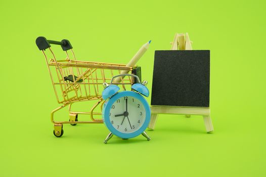 Shopping concept with shopping cart, alarm clock and small chalk board over a green background