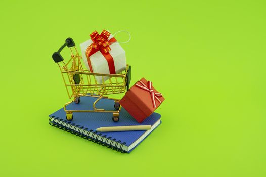 Shopping for gifts concept with open spiral bound notebook and shopping cart with gifts with decorative red ribbons and pencil over a green background with copy space