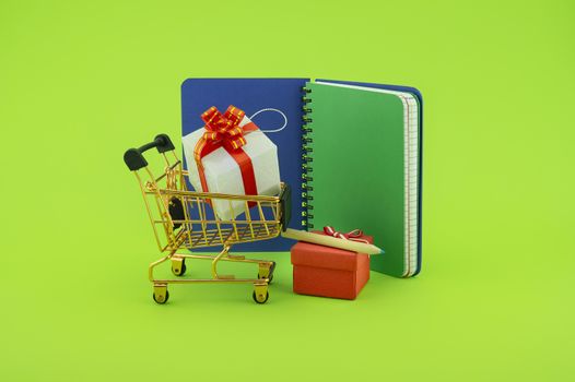 Shopping for gifts concept with open spiral bound notebook and shopping cart with gifts with decorative red ribbons and pencil over a green background with copy space