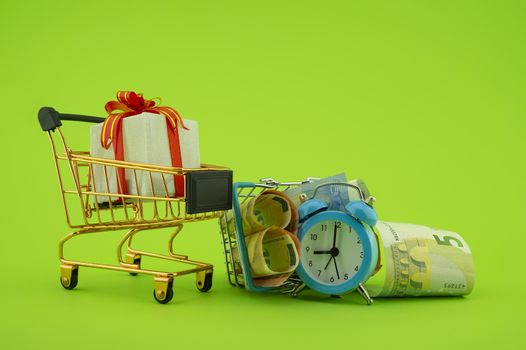 Shopping concept with shopping cart, small gift box, basket filled with cash banknotes and alarm clock over a green background