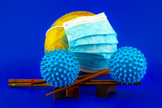 Coronavirus of Covid-19 pandemic still life with two blue virus molecules balanced on chopsticks with a surgical mask around world globe behind over a blue background