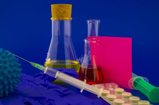 Concept of research for a virus vaccine with a conical lab flasks with yellow and red solutions alongside a hypodermic syringe and blue virus molecules with blank square of paper note for text