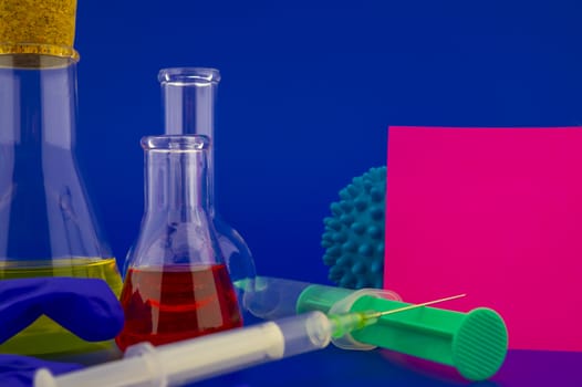 Concept of research for a virus vaccine with a conical lab flasks with yellow and red solutions alongside a hypodermic syringe and blue virus molecules with blank square of paper note for text