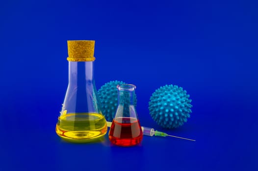 Concept of research for a Covid-19 vaccine with a conical lab flasks with yellow and red solutions alongside a hypodermic syringe and blue virus molecules over a blue background with copy space