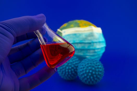 Coronavirus or Covid-19 still life with a gloved hand holding a laboratory flask with liquid or a possible vaccine in front of a globe with face mask and two virus molecules in blue toned theme