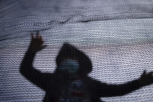 Silhouette of a man wearing hood and face masks for protection against coronavirus or COVID-19 behind a net or fence as concept for quarantine and social protest during isolation