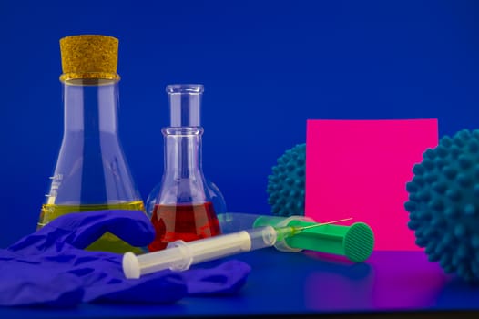 Concept of research for a virus vaccine with a conical lab flasks with yellow and red solutions alongside a hypodermic syringe and blue virus molecules with blank square of paper note for text