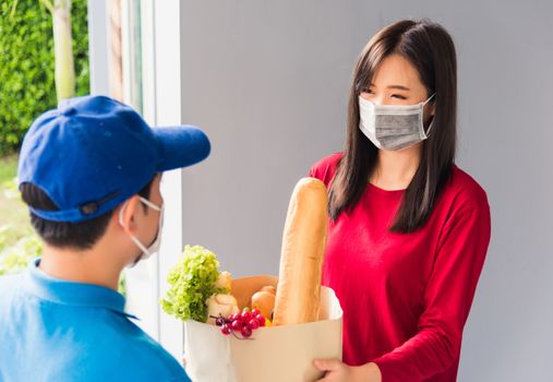 Asian young delivery man in uniform wear protective face mask he making grocery service giving fresh food to woman customer receiving front house under pandemic coronavirus, Back to new normal concept