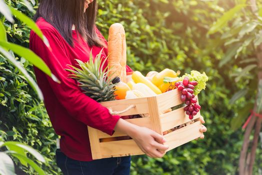 Portrait of Asian beautiful young woman farmer standing she smile and holding full fresh food raw vegetables fruit in a wood box in her hands on green leaves background