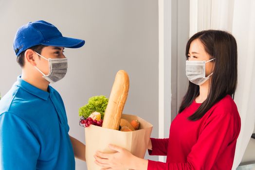 Asian young delivery man in uniform wear protective face mask he making grocery service giving fresh food to woman customer receiving front house under pandemic coronavirus, Back to new normal concept