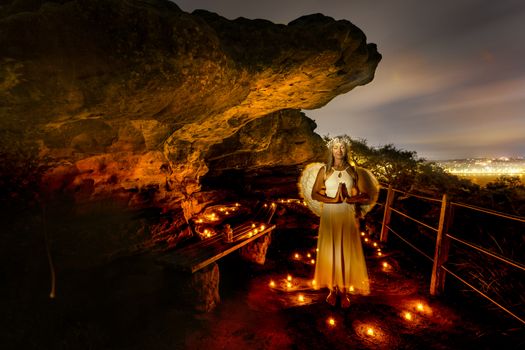 Praying angel surrounded by tea light candles at a small rock cave at night