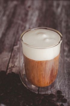 Coffee time and refreshment concept. Transparent glass of coffee with milk froth and roasted coffee bean on wooden background.