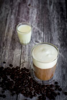 Coffee time and refreshment concept. Transparent glass of coffee with milk froth and roasted coffee bean on wooden background.