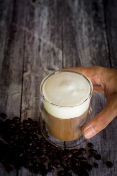 Coffee time and refreshment concept. Hand holding transparent glass of coffee with milk froth and roasted coffee bean on wooden background.