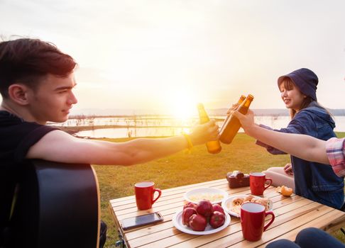 A group of Asian friends tourist drinking alcoholic beer and playing guitar together with happiness in Summer while having camping near lake at sunset