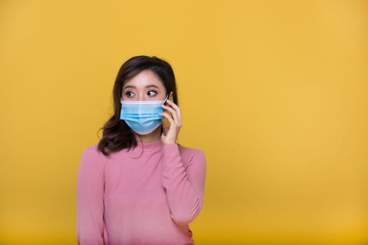 Portrait Asian beautiful happy young woman wearing  face mask or protective mask against coronavirus crisis or COVID-19 outbreak and she is using mobile phone or smartphone on yellow background