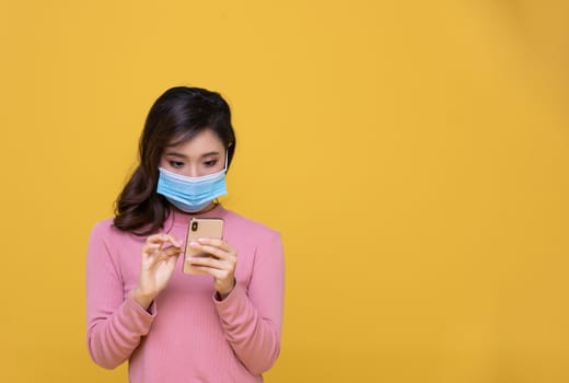 Portrait Asian beautiful happy young woman wearing  face mask or protective mask against coronavirus crisis or COVID-19 outbreak and she is using mobile phone or smartphone on yellow background