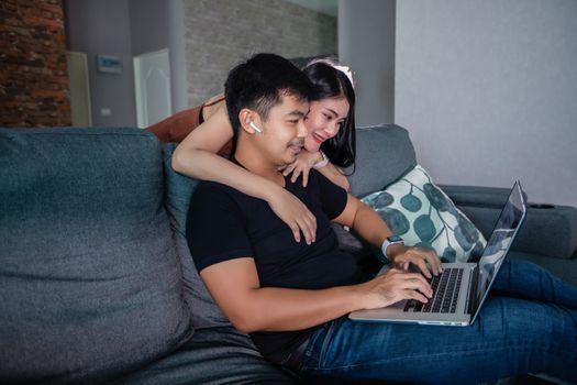 Asian young smiling couple hugging and using wireless earphones and using computer notebook working from home .He is prepare graphs and information for online meetings at home.