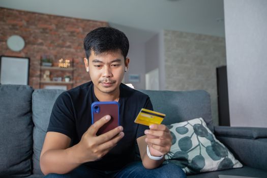 Asian man is holding credit card and using smart phone shopping online at home.Online shopping, e-commerce, internet banking, spending money, working from home concept