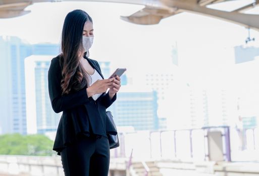 Business girl with hygiene mask stand on platform and use mobile phone and wait the sky train during go to work in situation of Covid-19 pandemic in city.