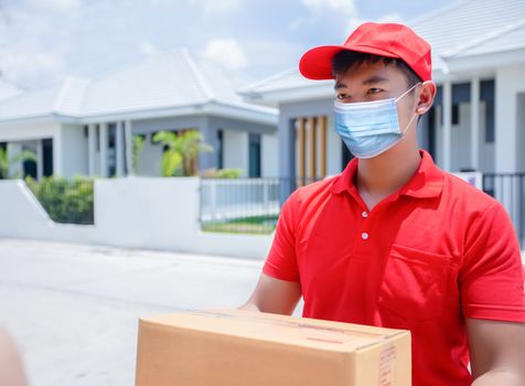 Asian delivery servicemen wearing a red uniform with a red cap and face mask handling cardboard boxes to give to the female customer in front of the house. Online shopping and Express delivery