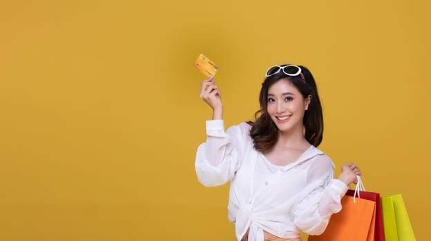 Portrait Asian beautiful happy young woman with sunglasses smiling cheerful and she is holding credit card and using smart phone for shopping online with shopping bags on yellow background.Happiness, consumerism, sale and people shopping concept
