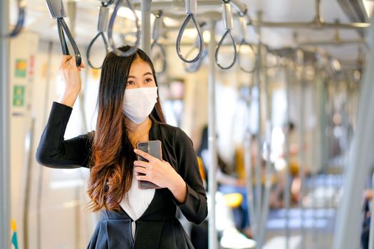 Portrait of beautiful business woman with hygiene mask hold mobile phone and stand with holding handrail in sky train also look to right side.