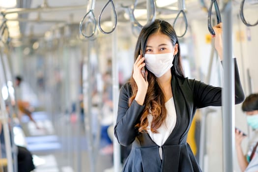 Portrait of beautiful business woman with hygiene mask use mobile phone and stand with hold handrail in sky train. She still work during pandemic of Covid-19 coronavirus spread in city.