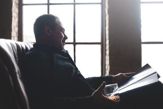 A relaxed businessman is drinking an expensive drink and reading a magazine.