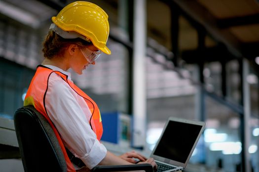 Pretty technician or engineer or worker sit on chair and use laptop in factory.