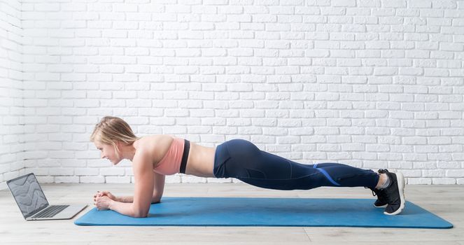 Stay home. Home fitness. Young blond woman doing plank at home looking at the laptop, white brick wall and wooden floor