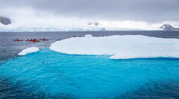 Huge iceberg with blue underwater part and small tourists kayaks with antarctic lagoon in the backgound, Peterman island, Antarctic peninsula