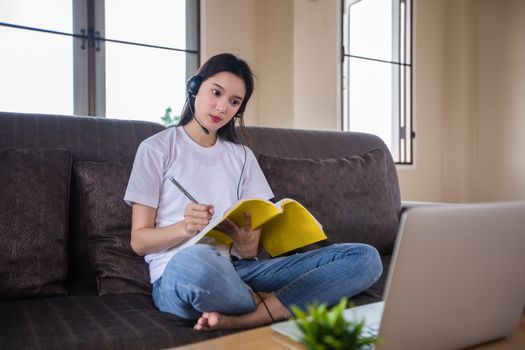 Asian happy woman student wear headphones study online watching webinar podcast on laptop listening learning education course conference calling and write notes look at laptop sit on sofa at home