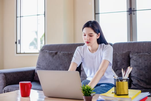 Asian woman pleased happy cheerful cute beautiful is using a notebook computer and sitting on sofa for working from home. She is quarantined at home during the coronavirus epidemic.