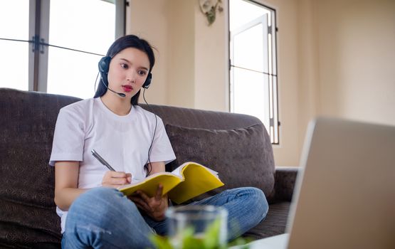 Asian happy woman student wear headphones study online watching webinar podcast on laptop listening learning education course conference calling and write notes look at laptop sit on sofa at home