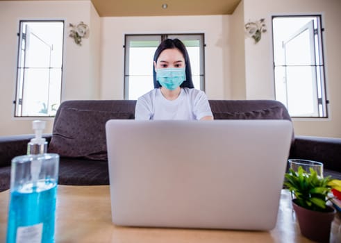 Asian woman cute beautiful is wearing protective mask and using a notebook computer and sitting on sofa for working from home. She is quarantined at home .And she has sanitizer gel on the table 