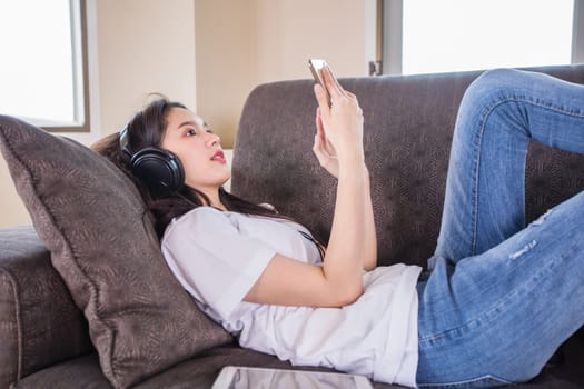 Asian young woman happy cheerful cute beautiful wears a headset for listening to music from a mobile phone and she is playing smartphone and lying on the sofa at home, 