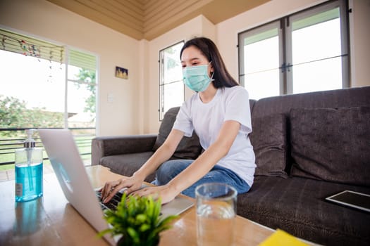 Asian woman cute beautiful is wearing protective mask and using a notebook computer and sitting on sofa for working from home. She is quarantined at home .And she has sanitizer gel on the table 