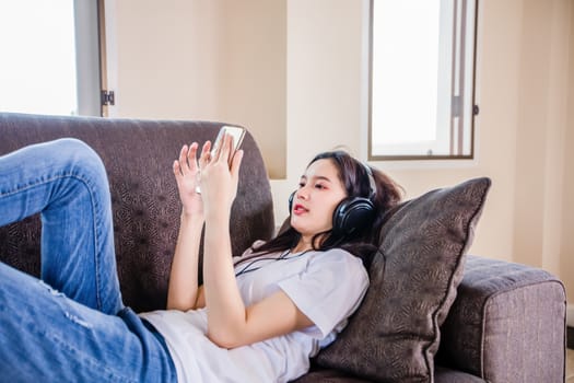Asian young woman happy cheerful cute beautiful wears a headset for listening to music from a mobile phone and she is playing smartphone and lying on the sofa at home, 