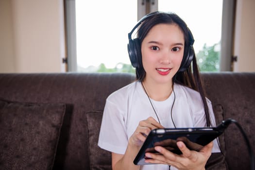 Asian young woman happy cheerful cute beautiful wears a headset for listening to music from a tablet and she is playing tablet and sitting on the sofa at home, 